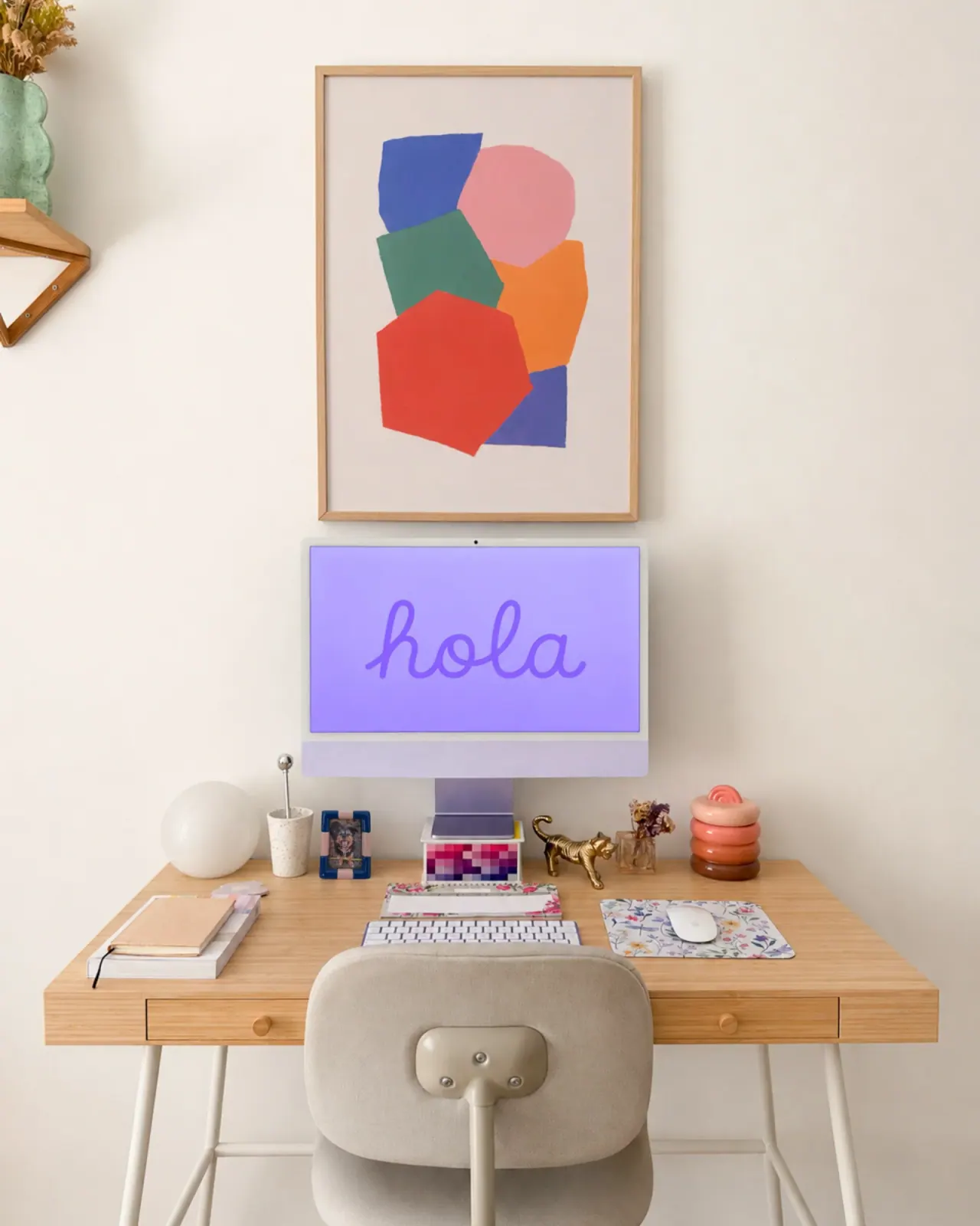 Escritorio de aprop social media con iMac lila mostrando un saludo en script, lámina abstracta colorida enmarcada y objetos personales sobre mesa de madera