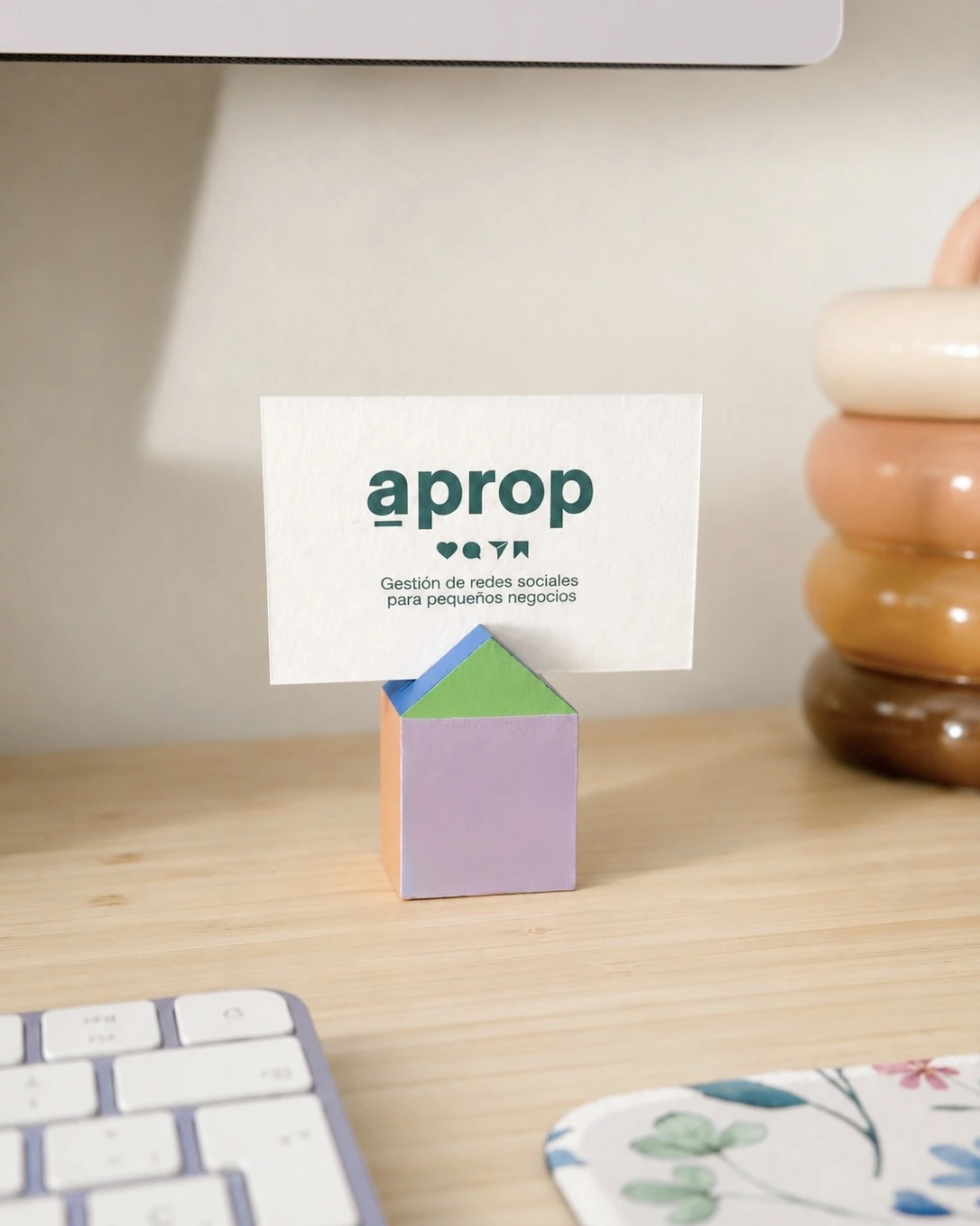 Tarjeta de aprop apoyada en un soporte con forma de casita de colores sobre escritorio de madera